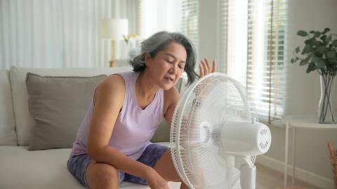 A middle-aged woman in a tank top and shorts sits in front of a white fan, trying to cool down.